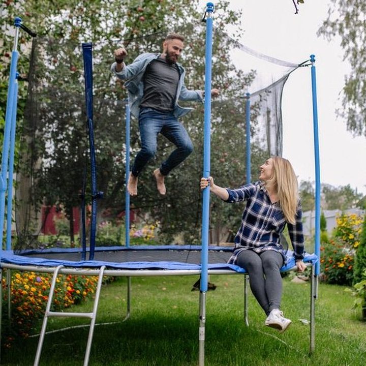 A man mid-jump on a garden trampoline while a woman sits on the edge laughing